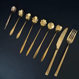 Puede incluir: Un juego de cubiertos dorados, que incluye un cuchillo, un tenedor y ocho cucharas. Las cucharas tienen diferentes diseños, incluyendo una cuchara redonda, una cuchara en forma de corazón y varias cucharas con cuencos en forma de flor.