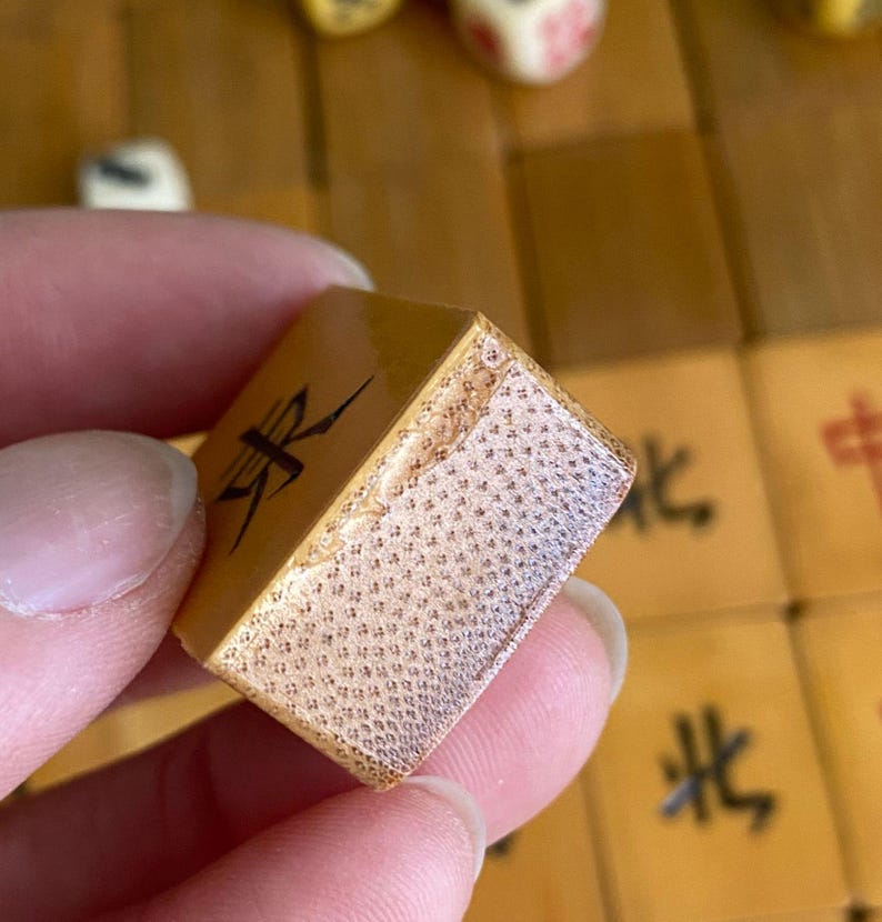 May include: A close-up of a tan Mahjong tile with black Chinese characters. The tile has a textured surface and is held by a person. Other tiles and dice are visible in the background.