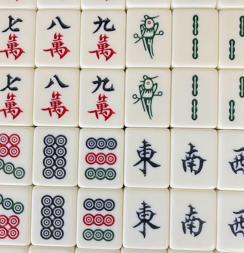 Puede incluir: Primer plano de una colecci&oacute;n de fichas de Mahjong blancas. Las fichas presentan varios s&iacute;mbolos y caracteres chinos en rojo, verde y negro. Algunas fichas muestran conjuntos de c&iacute;rculos, otras caracteres chinos.