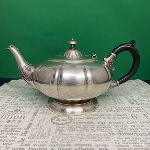 May include: A silver teapot with a black handle and a decorative spout. The teapot has a lid with a small finial and sits on a round base. The background is a green wall and a newspaper.