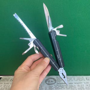 May include: A black multi-tool with silver metal implements including pliers, a serrated blade, and a knife. The handle features white printed markings labelled INCH and POCKET TOOL, with a ruler scale ranging from zero to three inches visible.