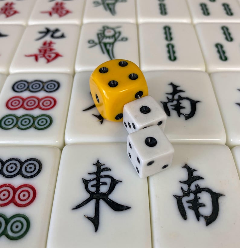 Puede incluir: Primer plano de un juego de Mahjong con fichas blancas con s&iacute;mbolos negros y rojos. Un dado amarillo y dos dados blancos est&aacute;n encima de las fichas. El juego est&aacute; destinado al uso recreativo.