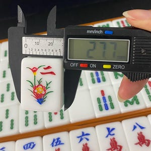 May include: A white Mahjong tile with a floral design, being measured with a digital caliper. The tile features a red and yellow flower with green leaves and blue accents. The caliper reads 27.7 mm, approximately 1.09 inches.
