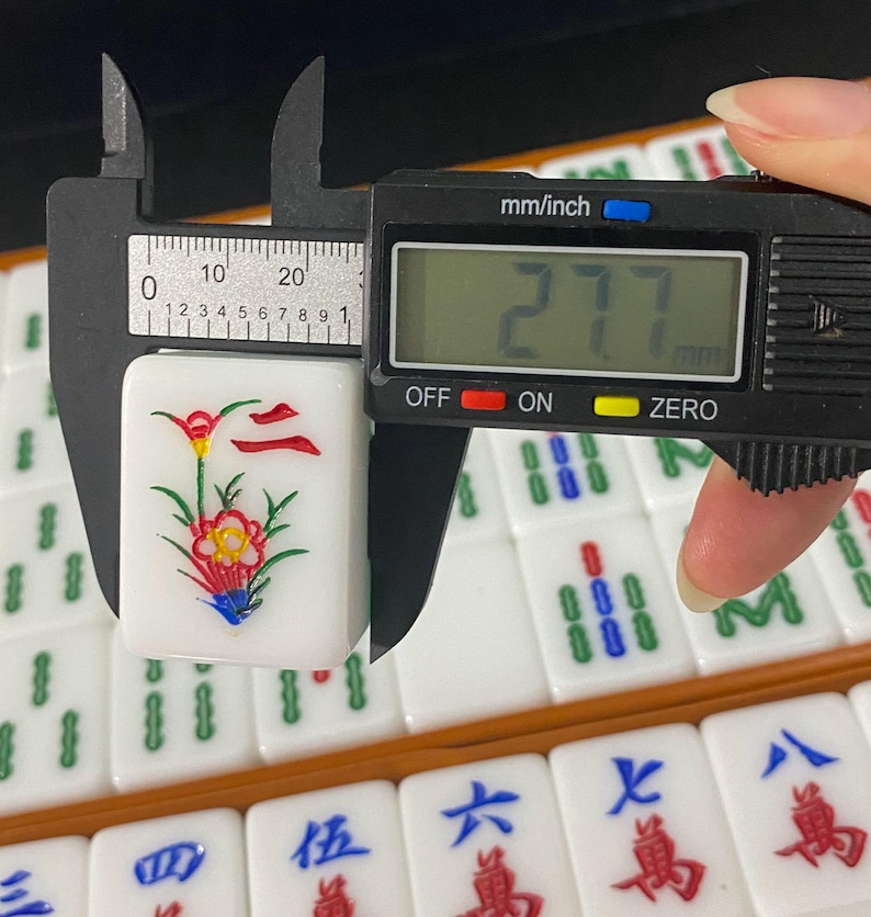 May include: A white Mahjong tile with a floral design, being measured with a digital caliper. The tile features a red and yellow flower with green leaves and blue accents. The caliper reads 27.7 mm, approximately 1.09 inches.