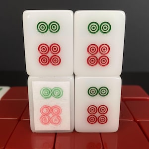 May include: Four white Mahjong tiles with green and red circular designs. The tiles are stacked, with the top two tiles showing two green circles above four red circles. The bottom two tiles mirror the design, with one tile partially obscured by a paper label.