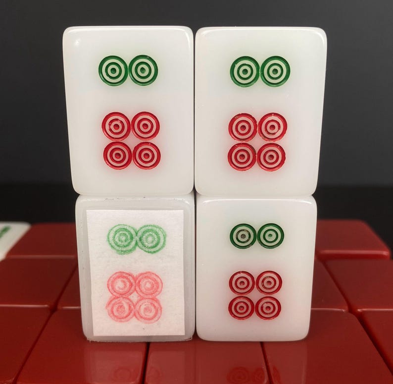 May include: Four white Mahjong tiles with green and red circular designs. The tiles are stacked, with the top two tiles showing two green circles above four red circles. The bottom two tiles mirror the design, with one tile partially obscured by a paper label.