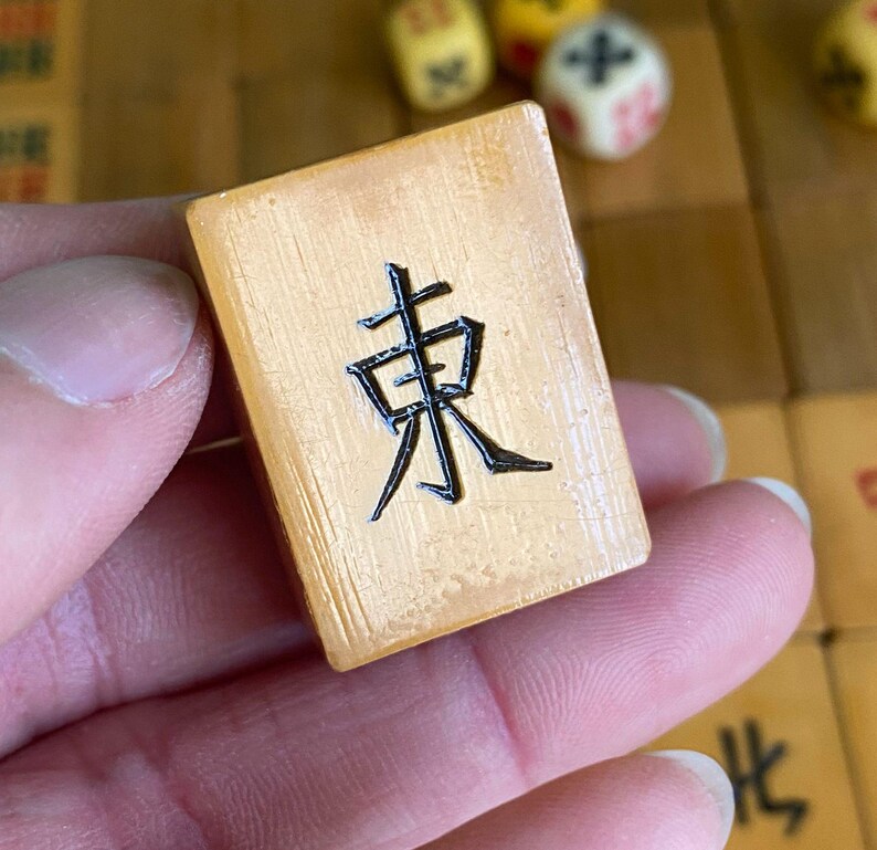 May include: A close-up of a Mahjong tile with the Chinese character for "East" in black. The tile is a light tan color and has a textured surface. Other Mahjong tiles and dice are visible in the background.