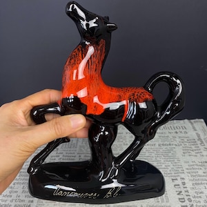 May include: A glossy black and red ceramic horse figurine. The horse is posed with its head raised and has a sleek, modern design. The base is black and has the inscription "Vancouver, B.C."