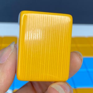 Puede incluir: Primer plano de una ficha de Mahjong amarilla. La ficha es rectangular con esquinas redondeadas y detalles acanalados verticales. La ficha es de color amarillo brillante y s&oacute;lido. El fondo est&aacute; desenfocado.