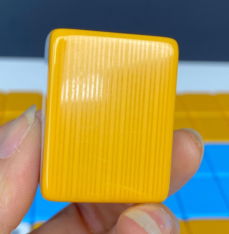 Puede incluir: Primer plano de una ficha de Mahjong amarilla. La ficha es rectangular con esquinas redondeadas y detalles acanalados verticales. La ficha es de color amarillo brillante y s&oacute;lido. El fondo est&aacute; desenfocado.