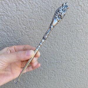 Peut inclure: Un chausse-pied en argent avec un motif floral sur la poignée. La poignée est incurvée et le crochet se trouve à l'extrémité d'un long manche fin.