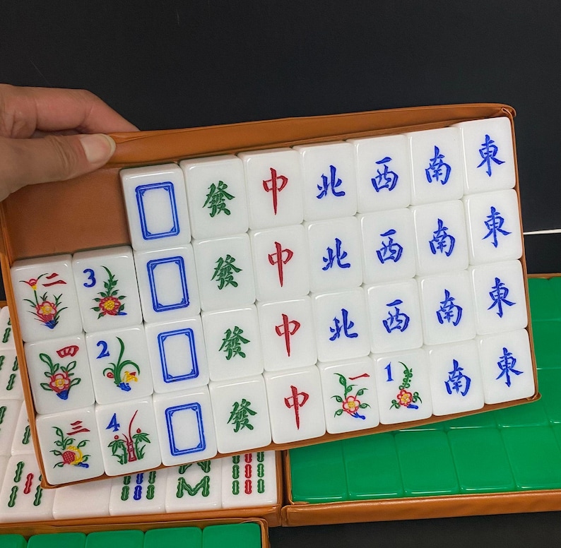 May include: A collection of white Mahjong tiles, each bearing Chinese characters and symbols, presented in a brown case. The tiles showcase diverse designs, including floral patterns, numbers, and directional markers. Some tiles are visible, while others are in the background.