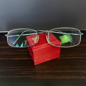 Puede incluir: Gafas con montura azul claro y lentes rectangulares sobre una pequeña caja roja. Las gafas tienen un puente y patillas plateadas. La caja es de color rojo liso con un patrón decorativo alrededor de la parte superior.