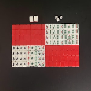 Puede incluir: Un juego de Mahjong completo con fichas rojas y blancas, dispuestas sobre una superficie negra. Las fichas muestran caracteres y s&iacute;mbolos chinos. Dados y fichas de repuesto tambi&eacute;n est&aacute;n presentes.