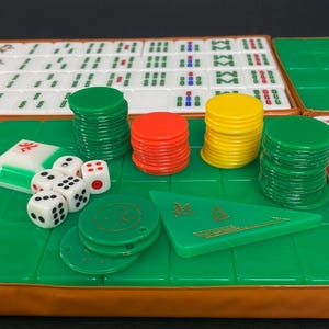 May include: A Mahjong set featuring white tiles, green, red, and yellow chips, dice, and a green triangular piece. The set is displayed on a green surface with a brown border, ready for play.