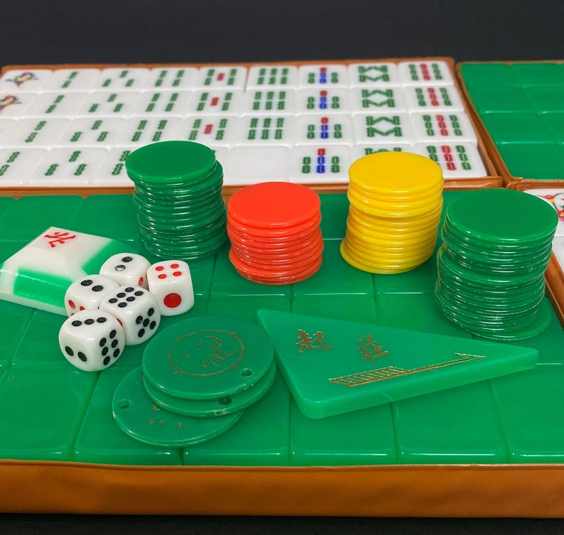 May include: A Mahjong set featuring white tiles, green, red, and yellow chips, dice, and a green triangular piece. The set is displayed on a green surface with a brown border, ready for play.