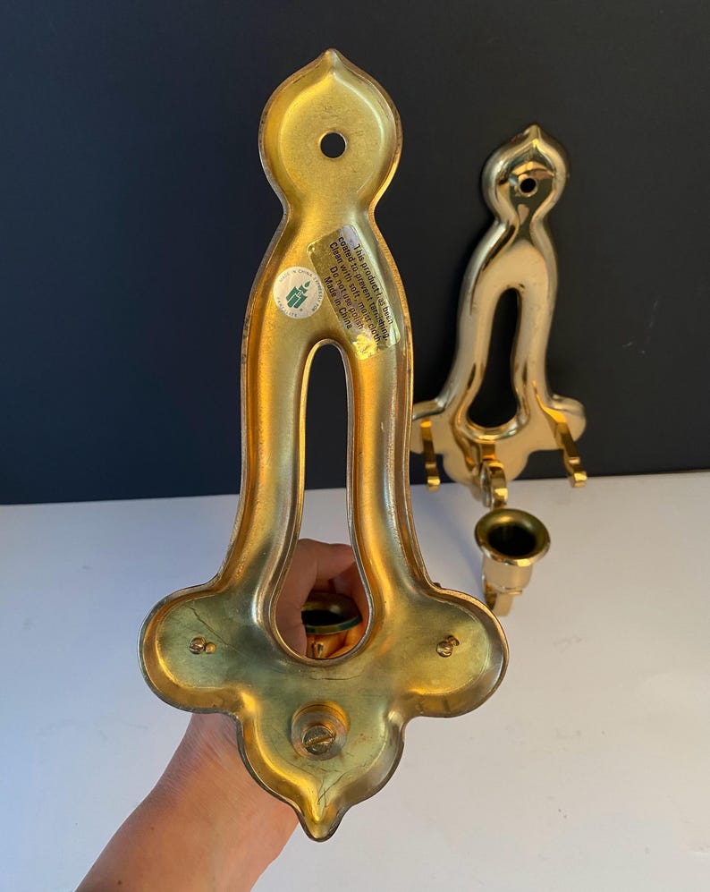 May include: Two vintage brass candle holders. The candle holders have a decorative, ornate design with a curved shape and a small cup for the candle. The brass has a shiny, polished finish. The text on the sticker says "This product is coated to prevent tarnishing. Clean with soft, mold cloth. Made in China."