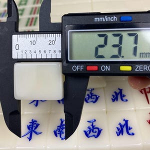 May include: A white Mahjong tile is measured with a digital caliper. The caliper displays a measurement of 23.7 mm. The tile is rectangular and sits on a surface of other Mahjong tiles with blue and red characters.