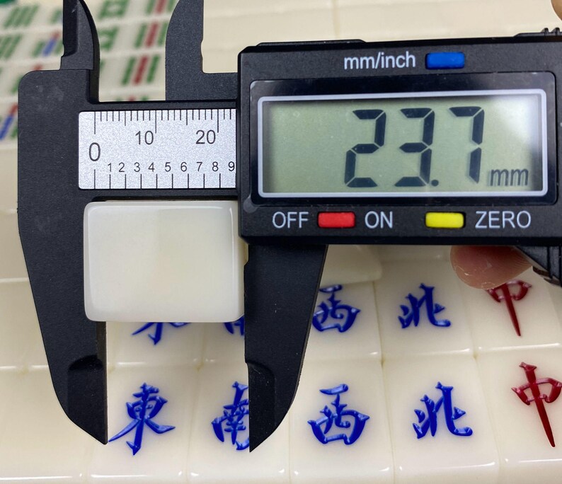 May include: A white Mahjong tile is measured with a digital caliper. The caliper displays a measurement of 23.7 mm. The tile is rectangular and sits on a surface of other Mahjong tiles with blue and red characters.