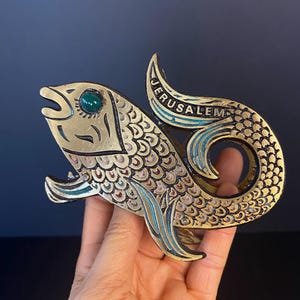 May include: A brass fish sculpture with a green gem eye and the word "JERUSALEM" in blue lettering. The fish has detailed scale patterns and blue and gold accents. The sculpture is held in a hand.