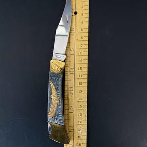 May include: A stainless steel folding knife with a silver blade and a handle featuring a gold eagle design. The knife is approximately 6 inches long and is next to a wooden ruler.