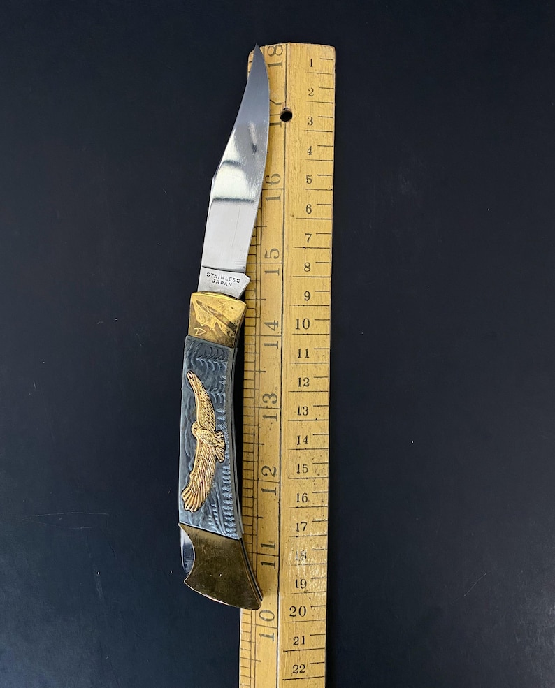 May include: A stainless steel folding knife with a silver blade and a handle featuring a gold eagle design. The knife is approximately 6 inches long and is next to a wooden ruler.