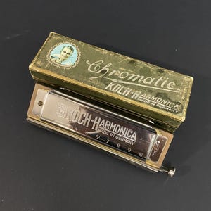 Könnte beinhalten: Eine silberfarbene Koch-Harmonika aus Metall mit nummerierten Löchern von 4 bis 10, zusammen mit der originalen grünen Pappschachtel. Die Schachtel trägt die Aufschrift "Chromatic", "Koch-Harmonica" und "Made in Germany" sowie ein rundes Porträt.