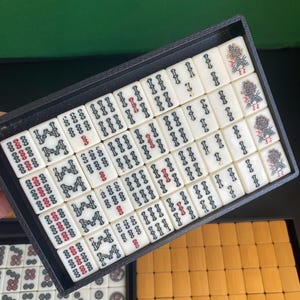 May include: A black tray filled with ivory-colored Mahjong tiles. The tiles feature black and red symbols, with some displaying floral designs. The tray is set against a green background, with a glimpse of additional tiles in a separate tray.