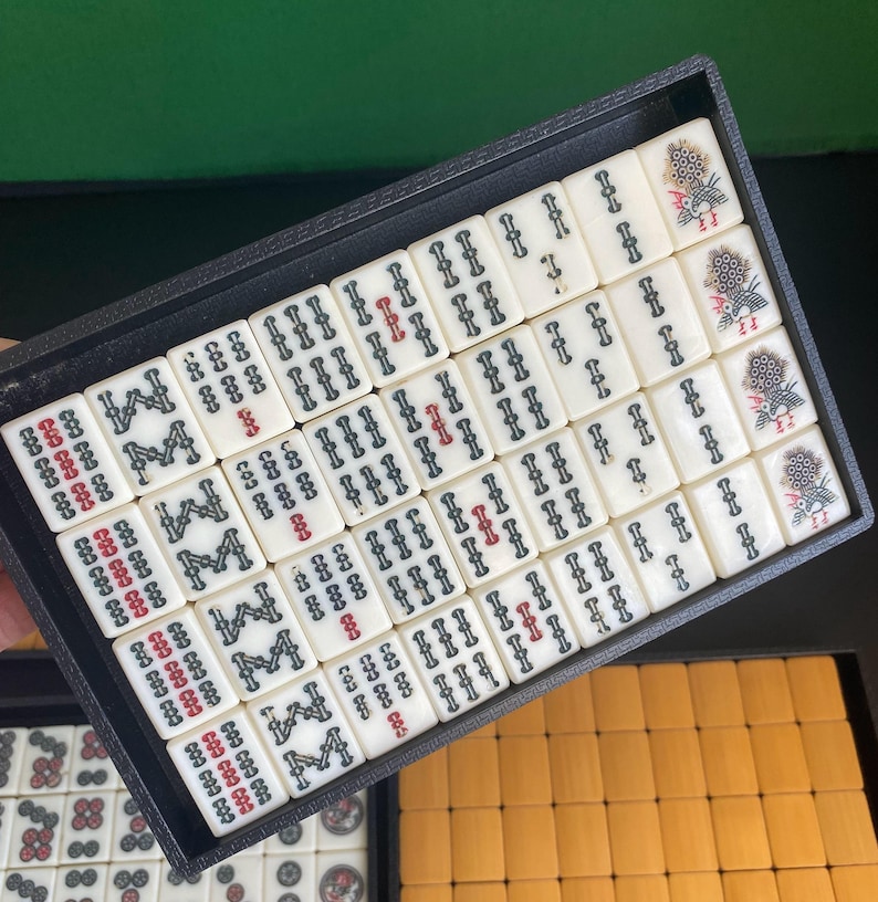 May include: A black tray filled with ivory-colored Mahjong tiles. The tiles feature black and red symbols, with some displaying floral designs. The tray is set against a green background, with a glimpse of additional tiles in a separate tray.