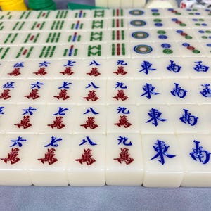 May include: A close-up of a Mahjong set with ivory-colored tiles. The tiles feature red and blue Chinese characters and symbols. The set includes green, yellow, and white game pieces.