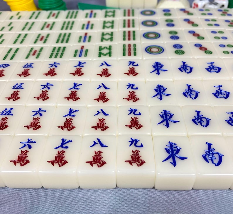 May include: A close-up of a Mahjong set with ivory-colored tiles. The tiles feature red and blue Chinese characters and symbols. The set includes green, yellow, and white game pieces.