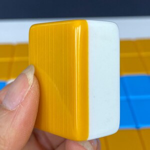 Puede incluir: Primer plano de una ficha de Mahjong, sostenida en una mano. La ficha es rectangular, con una superficie amarilla brillante, con estr&iacute;as verticales en un lado y una superficie blanca lisa en el otro. El fondo muestra otras fichas en amarillo, azul y blanco.