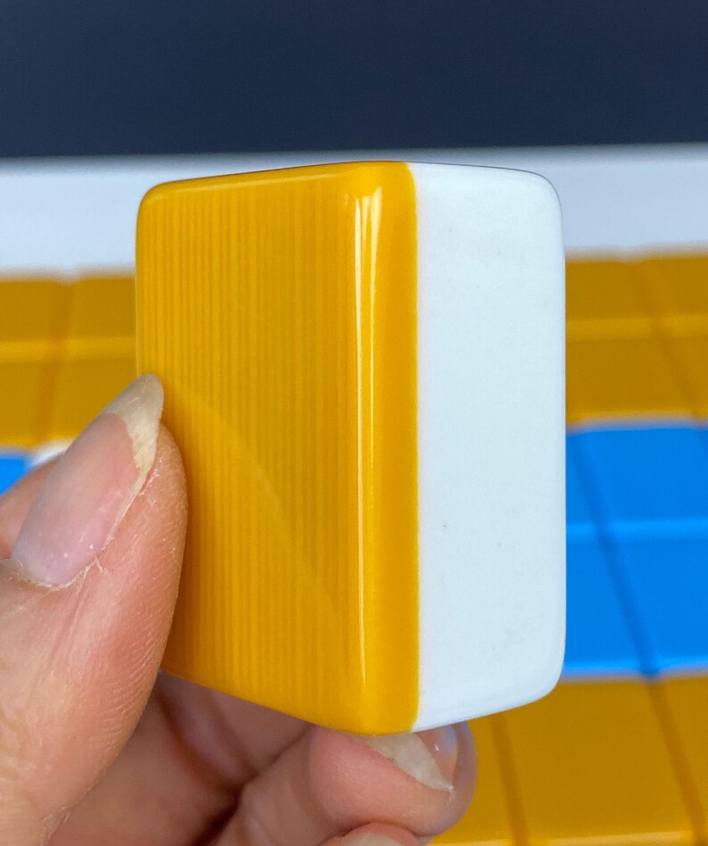 Puede incluir: Primer plano de una ficha de Mahjong, sostenida en una mano. La ficha es rectangular, con una superficie amarilla brillante, con estr&iacute;as verticales en un lado y una superficie blanca lisa en el otro. El fondo muestra otras fichas en amarillo, azul y blanco.