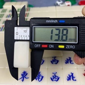 May include: A white rectangular Mahjong tile is measured with a digital caliper. The caliper displays a measurement of 13.8 mm. Other Mahjong tiles with blue Chinese characters are visible in the background.