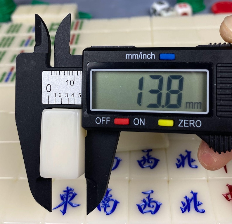 May include: A white rectangular Mahjong tile is measured with a digital caliper. The caliper displays a measurement of 13.8 mm. Other Mahjong tiles with blue Chinese characters are visible in the background.