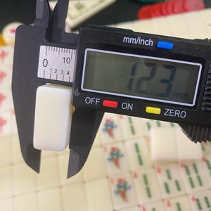 May include: A digital caliper measuring a cream-colored Mahjong tile. The tile is 12.3 mm wide, as indicated on the digital display. The caliper is black with a digital display showing the measurement in millimeters and inches.