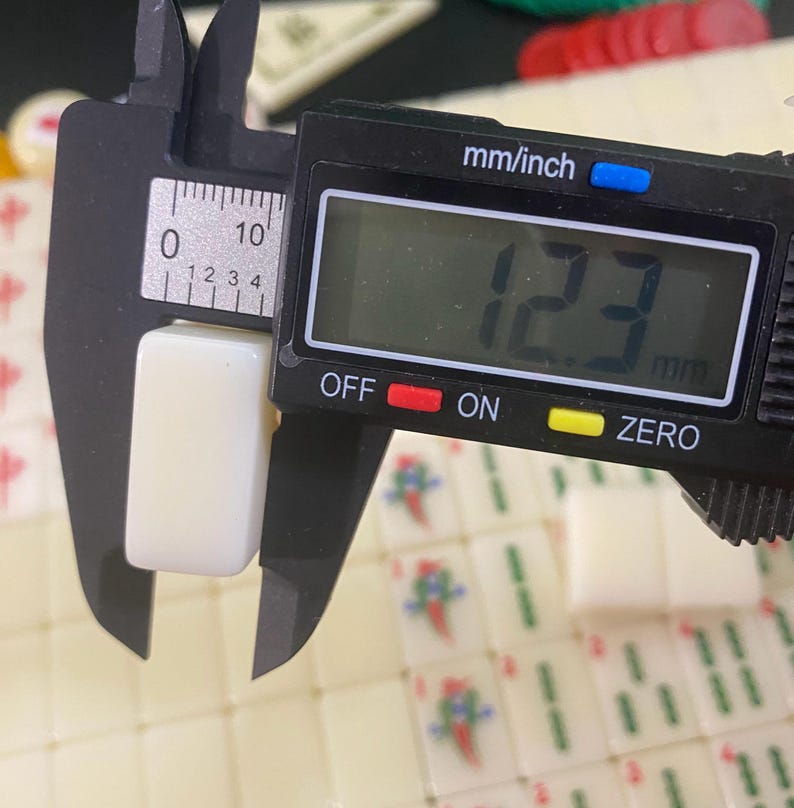 May include: A digital caliper measuring a cream-colored Mahjong tile. The tile is 12.3 mm wide, as indicated on the digital display. The caliper is black with a digital display showing the measurement in millimeters and inches.