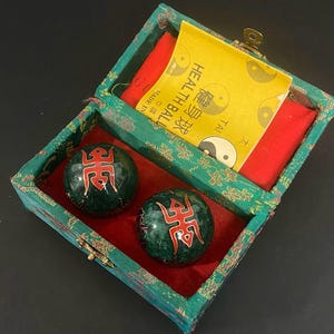 Peut inclure: Deux boules de santé sphériques vert foncé avec des symboles décoratifs rouges, rangées dans une boîte rectangulaire recouverte de tissu vert avec une doublure intérieure rouge. Une carte jaune avec le texte HEAL TH BALL et des caractères chinois.