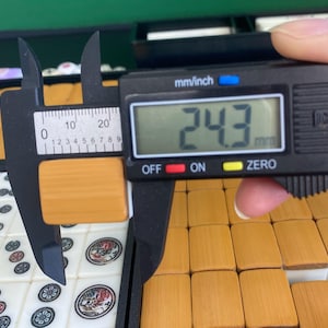 May include: A digital caliper measuring a rectangular, light brown tile. The digital display shows 24.3 mm. The tile is part of a set of similar tiles, some of which are visible in the background. The caliper is black with white markings.