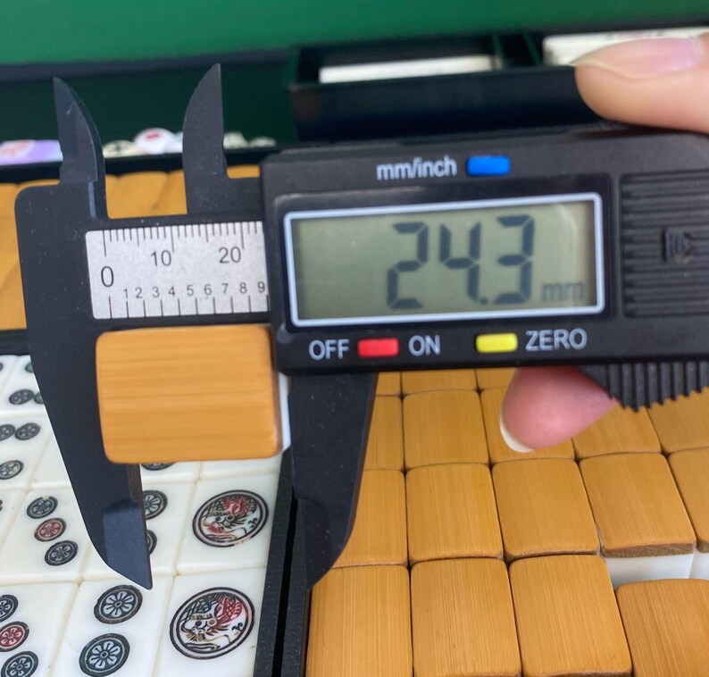 May include: A digital caliper measuring a rectangular, light brown tile. The digital display shows 24.3 mm. The tile is part of a set of similar tiles, some of which are visible in the background. The caliper is black with white markings.