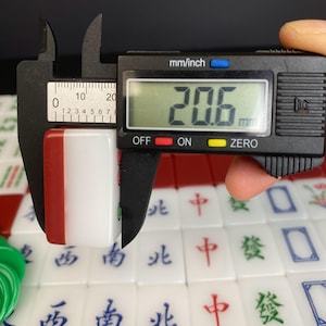 May include: A white and red Mahjong tile is measured with a digital caliper, showing a width of 20.6 mm. The tile has Chinese characters and symbols. Other tiles are visible in the background.
