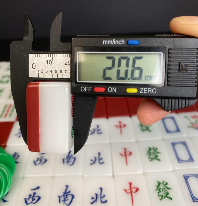 May include: A white and red Mahjong tile is measured with a digital caliper, showing a width of 20.6 mm. The tile has Chinese characters and symbols. Other tiles are visible in the background.