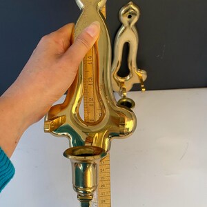 May include: Two brass wall sconces. The larger sconce is approximately 14 inches tall, with a candle cup and ornate design. The smaller sconce has a simpler design. A wooden ruler is held next to the larger sconce.