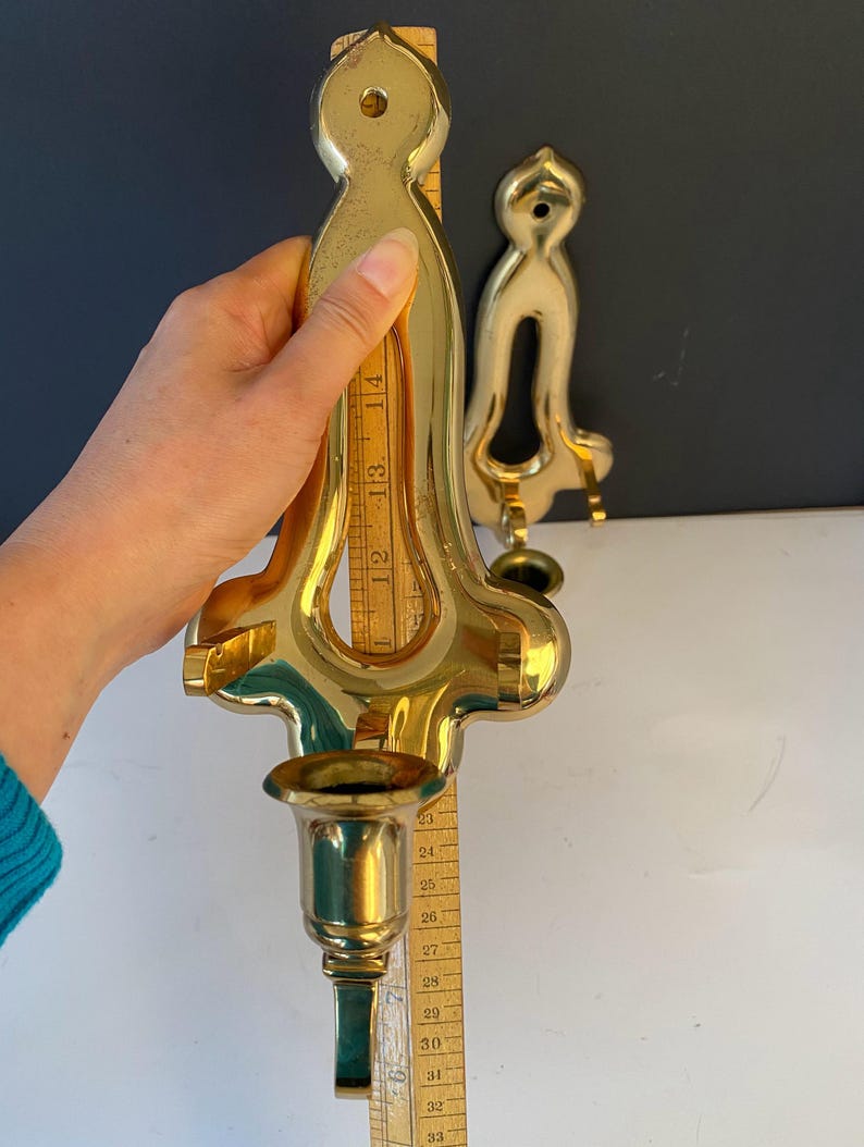 May include: Two brass wall sconces. The larger sconce is approximately 14 inches tall, with a candle cup and ornate design. The smaller sconce has a simpler design. A wooden ruler is held next to the larger sconce.