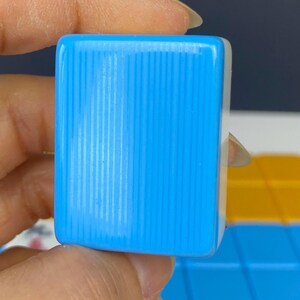 Puede incluir: Primer plano de una baldosa rectangular azul claro con l&iacute;neas verticales. La baldosa se sostiene entre los dedos. El fondo est&aacute; desenfocado, con toques de amarillo y azul.
