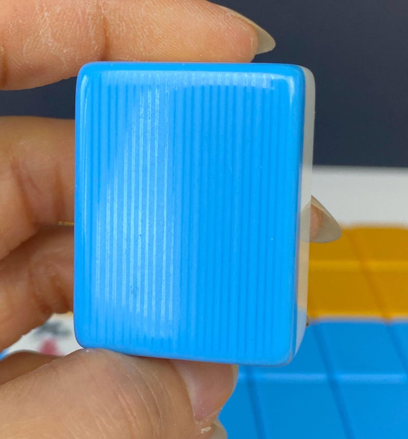 Puede incluir: Primer plano de una baldosa rectangular azul claro con l&iacute;neas verticales. La baldosa se sostiene entre los dedos. El fondo est&aacute; desenfocado, con toques de amarillo y azul.