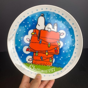 May include: A decorative plate featuring Snoopy in his doghouse decorated with Christmas lights. The plate has a blue background with white snowflakes and a white border with blue dots. The words "Christmas 1977" are written on the plate.