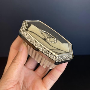 Könnte beinhalten: Eine silberfarbene Bürste mit rechteckigem, abgeschrägtem Design. Die Bürste hat einen dekorativen Rand und ein zentrales Emblem. Die Borsten sind weiß. Die Bürste wird in einer Hand vor dunklem Hintergrund gehalten.