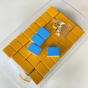 Puede incluir: Un recipiente de pl&aacute;stico transparente lleno de fichas de Mahjong amarillas y azules. Las fichas est&aacute;n dispuestas en un patr&oacute;n de cuadr&iacute;cula. Dos dados son visibles en una peque&ntilde;a bolsa de pl&aacute;stico. Las piezas del juego est&aacute;n sobre un fondo blanco.