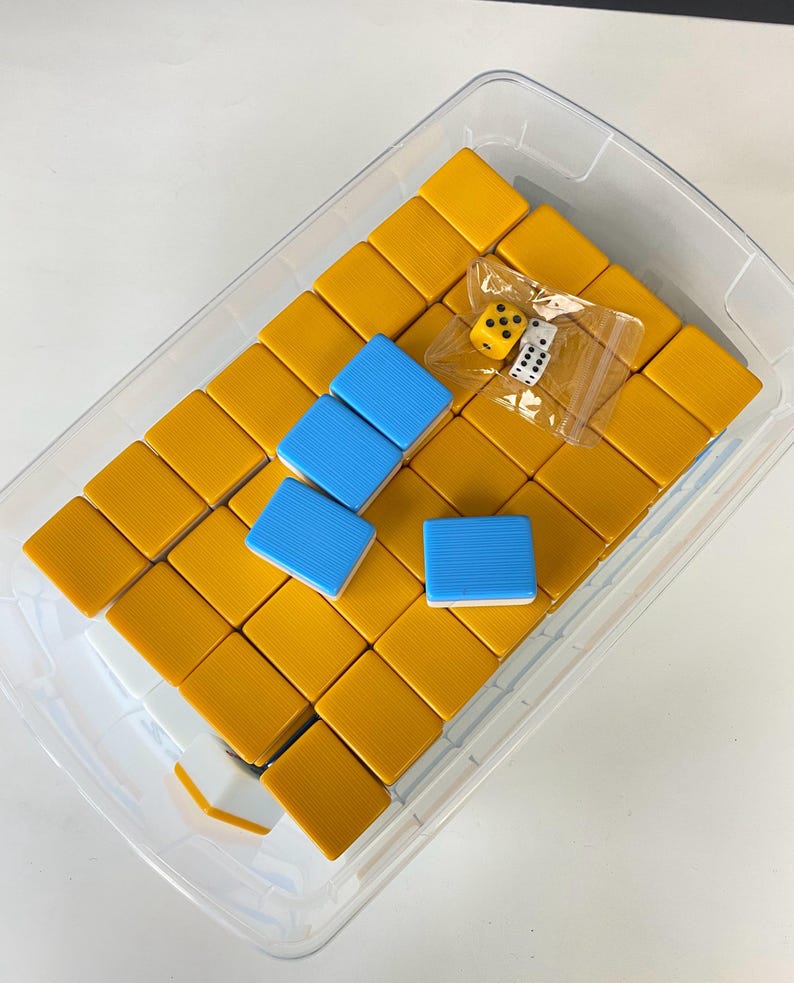 Puede incluir: Un recipiente de pl&aacute;stico transparente lleno de fichas de Mahjong amarillas y azules. Las fichas est&aacute;n dispuestas en un patr&oacute;n de cuadr&iacute;cula. Dos dados son visibles en una peque&ntilde;a bolsa de pl&aacute;stico. Las piezas del juego est&aacute;n sobre un fondo blanco.